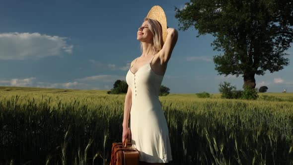 Woman in white dress with hat and suitcase in wheat field