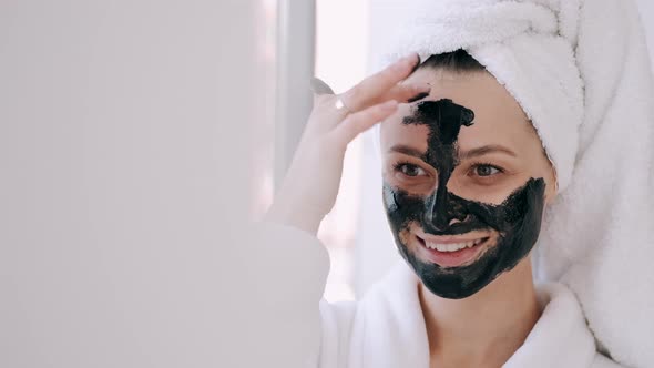 Pretty Woman with Wrapped Hair in a Towel Is Applying Black Mask on Face