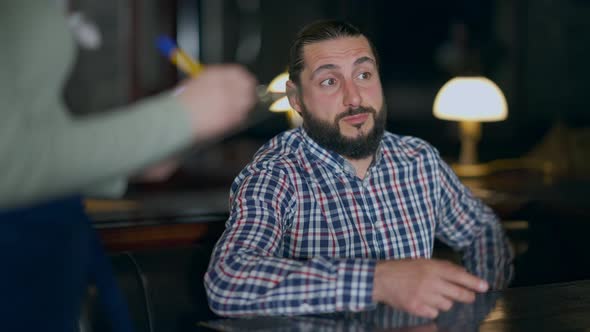 Portrait of Bearded Man Ordering Sitting at Table in Restaurant with Blurred Waitress Writing Order