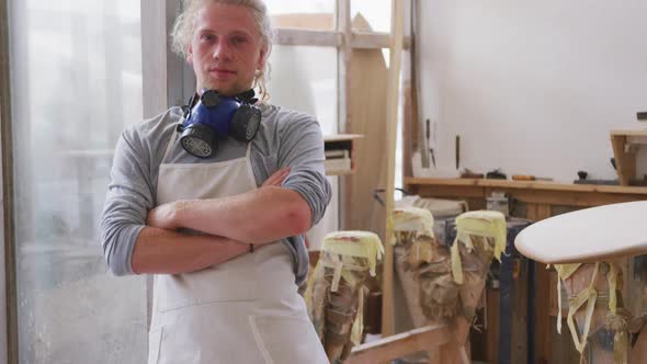 Caucasian male surfboard maker wearing a face mask and standing in his studio with his arms crossed