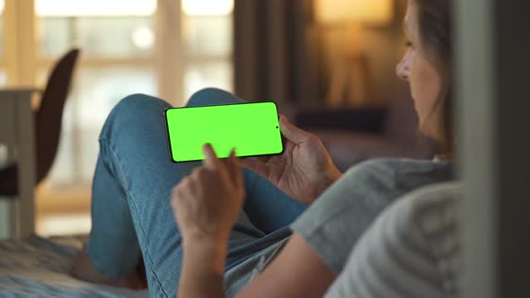 Woman at Home Lying on a Sofa and Using Smartphone with Green Mock-up Screen in Horizontal Mode