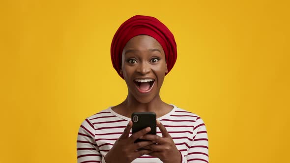 Excited Black Woman Holding Smartphone Texting Wearing Headwrap Yellow Background