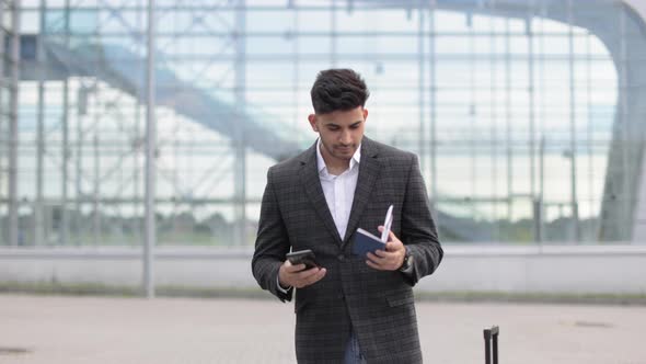 Likable Young Indian Man Using Mobile Phone Standing Outside Airport Terminal Holding Passport