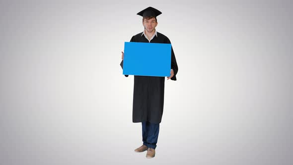 Smiling Graduate Male in Mortarboard Holding Blank Sign and Looking at It on Gradient Background