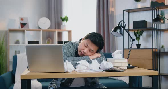 Asian Man Sleeping on the Office Table Covered with Many Crumpled Papers in Contemporary Home Office