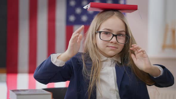 Intelligent Cute Schoolgirl Posing with Book on Head Sitting at Desk in School Classroom with