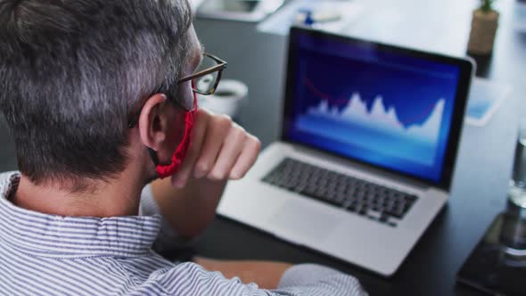 Caucasian man wearing face mask using laptop while sitting on his desk at modern office