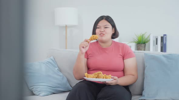 Asian oversize big women eat unhealthy food fried chicken on sofa while watching television in house