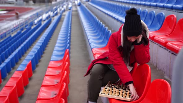 Young Woman in Winter Clothes Plays Chess Sitting on Stadium Bleachers Alone