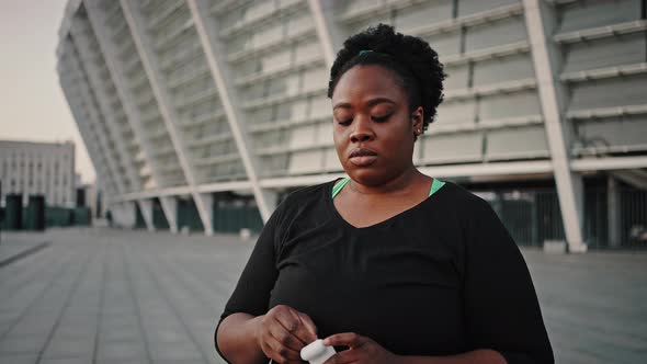 Young Overweight African American Woman Putting in Wireless Earphones Preparing for Walk in Evening