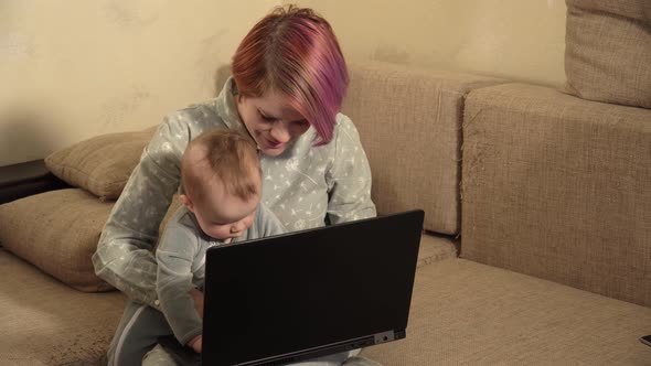 Young Woman Sitting on Sofa with Her Little Son on Knees and Typing on Laptop