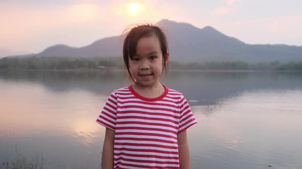 Cute little girl smiles and looks at the camera during the sunset by the lake.