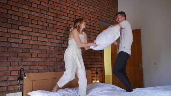 Wide Shot Pillow Fight of Joyful Caucasian Young Couple Standing on Bed