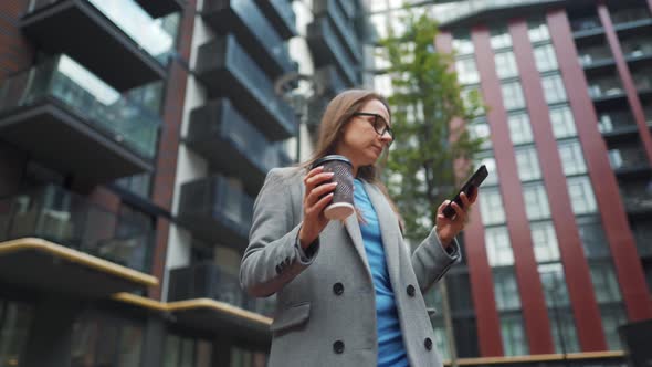 Formally Dressed Woman Walking Down the Street in a Business District with Coffee in Hand and Using