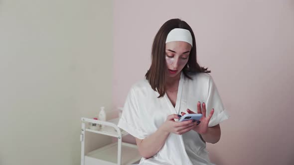 Young Woman with Collagen Pads Under Eyes Uses Her Phone in Beauty Clinic Salon
