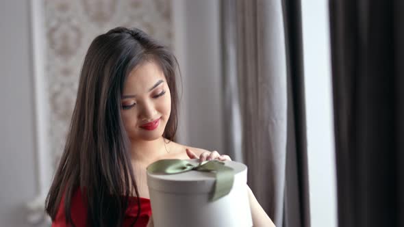 Happy Amazed Elegant Asian Woman Opening Gift Box and Smiling at Home Cozy Interior