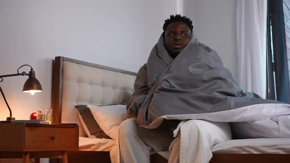 Unwell African American Young Man Shivering Sitting on Bed Wrapped in Blanket