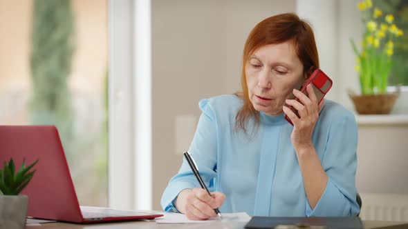 Busy Serious Mature Woman Talking on Phone Writing with Pen Surfing Internet on Laptop