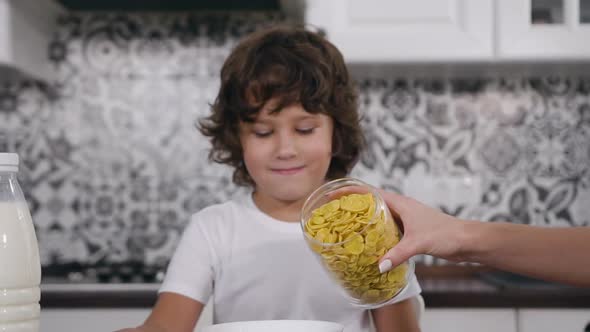 Little Boy Sitting at the Table and Waiting while His Mother Filling the Bowl with Corn Flakes