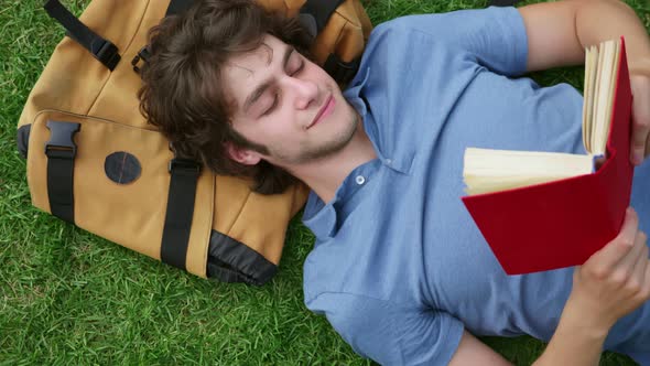 Top View of Young Man Lying on Grass Reading Book with Head on Backpack