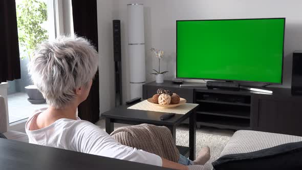 A Happy Middle-aged Woman Sits on a Sofa and Watches TV with a Green Screen, Then Turns and Smiles