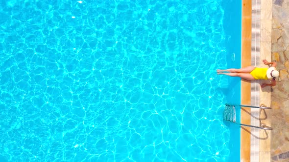 Aerial View of a Woman in Yellow Swimsuit and a Hat Sits on the Edge of the Pool