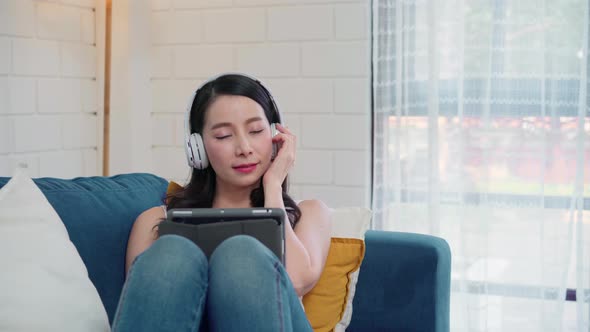 Asian woman listening music and using tablet, female using relax time lying on home sofa.