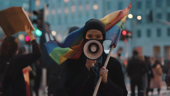 Woman with Face Mask Speaking Into the Megaphone While Holding Rainbow Flag. Demonstration Against