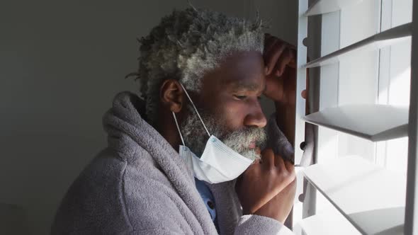 Senior man with face mask on his chin looking out of window at home