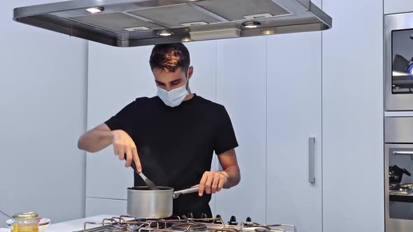 Young man is cooking food for delivery in his restaurant in protective mask.