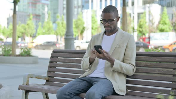 African Man Browsing Internet on Smartphone While Sitting Outdoor on Bench