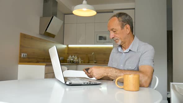 Trendy Mature Man is Working From Home with Laptop Sitting at the Table in His Kitchen Buying