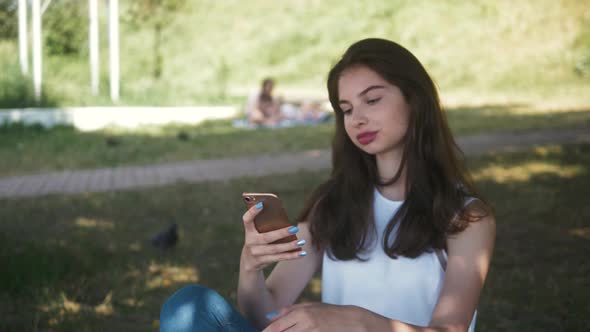 Beautiful Cheerful Girl with a Smartphone Sitting in a Park on a Bench on a Sunny Day Teenager