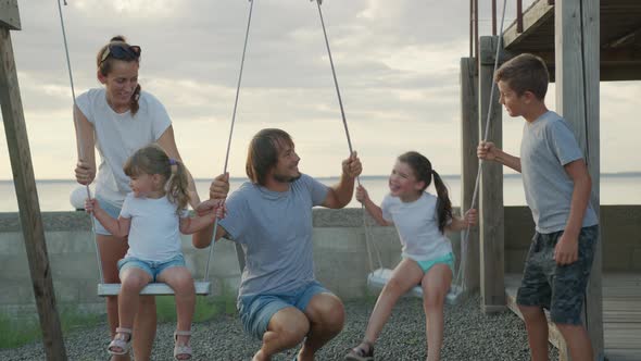 Happy Family Rolls Young Children on a Swing Outdoors at Sunset