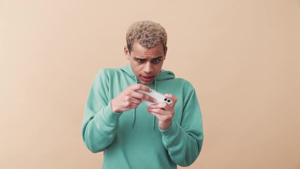 Happy curly-haired man wearing blue hoodie playing digital game by phone