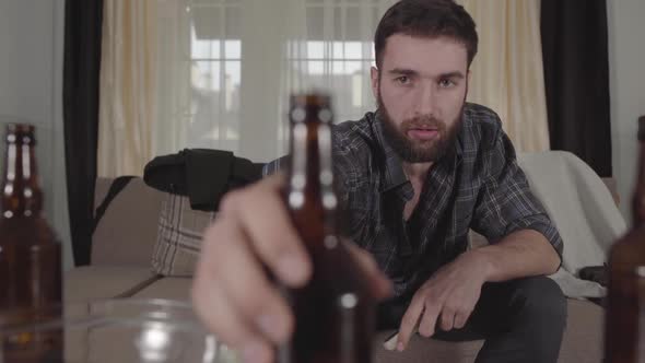 Young Bearded Man Sitting on the Sofa Takes Empty Beer Bottle From the Table and Look at It with