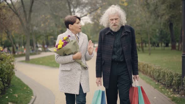 Confident Senior Caucasian Wife Walking with Husband in Autumn Park with Bouquet of Flowers and