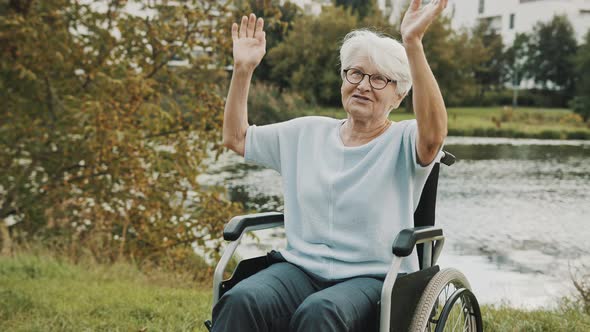 Disabled Senior Woman Dancing with Hands in the Wheelchair Near the River on the Autumn Day