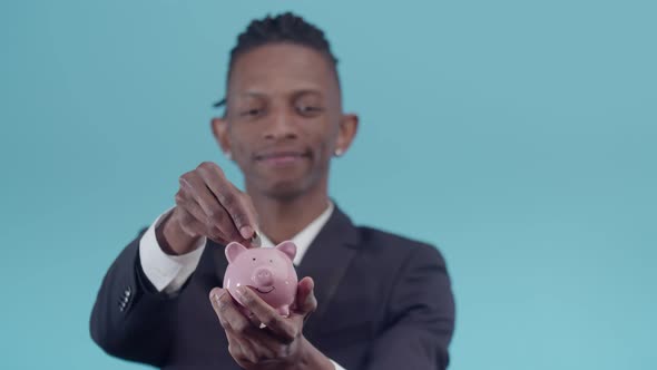 Black man in a strict suit looking into the camera puts coins in a piggy bank on blue background.