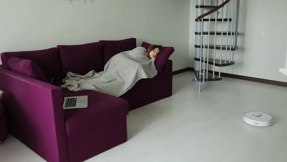 Free from household chores woman is relaxing on couch while robot vacuum cleaner cleanin. 