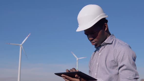 An AfricanAmerican Electrician in a Helmet Stands Against the Backdrop of a Windmill at an Air Power