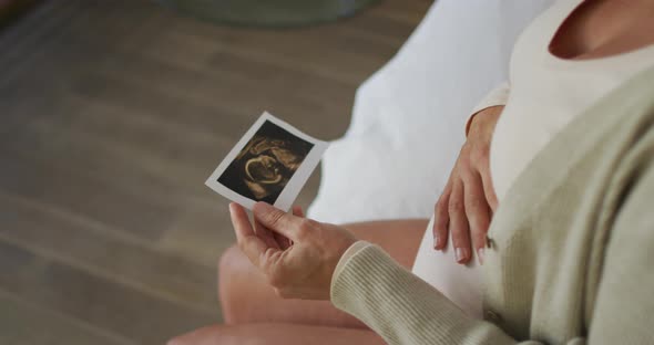 Hands of caucasian pregnant woman sitting on bed, touching belly and looking on ustrasound photo