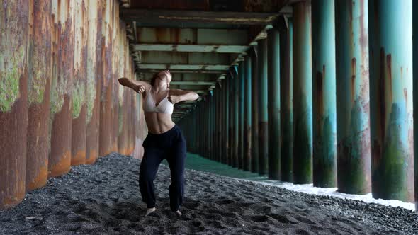 a Barefoot Woman Dances Under the Pillars of the Bridge Against the Background of the Incoming Waves