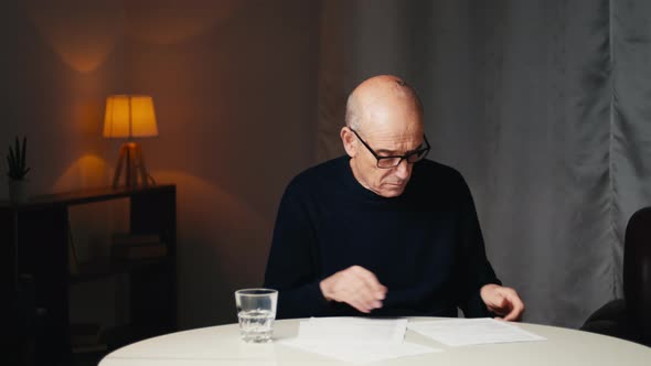 Elderly Man Sitting at the Table Reads His Electricity and Gas Bills