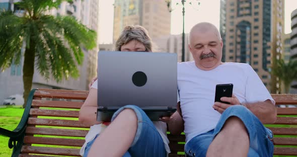 Happy Senior Couple Sitting on a Bench in the Summer in a Modern City with a Laptop