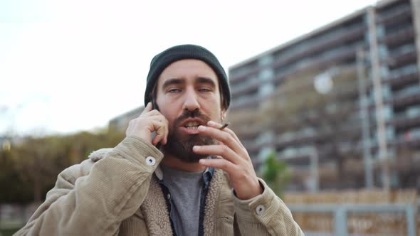 Smiling bearded man wearing hat talking by phone and smoking