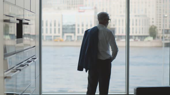 Back View of Mature Entrepreneur Looking Through Panoramic Window in Kitchen Putting on Jacket and