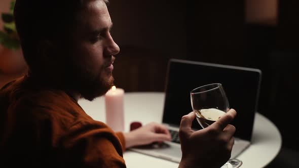 Closeup View of Thoughtful Young Man Drinking Alcohol From Glass During Working on Laptop Computer