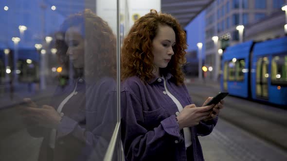 Pretty woman leaning on glass pane at train station using smartphone