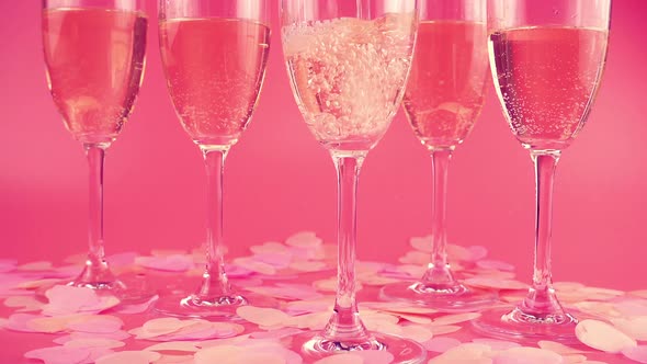 Pouring sparkling wine into glasses on a pink background with heart-shaped confetti. Slow motion.